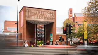 The exterior of The British Library, a sandstone building with "The British Library" carved above its entrance, with a metal gate moulded to say "British Library" repeatedly beneath it. Some people, blurred due to long exposure, walk in front of the library