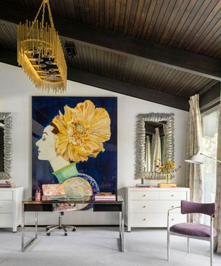 Dark wooden paneled ceiling, white painted walls, decorated with large colorful artwork, mirrors, two white storage chests, glass and black metal desk, purple upholstered dining chair, light gray carpet, bright gold modern chandelier