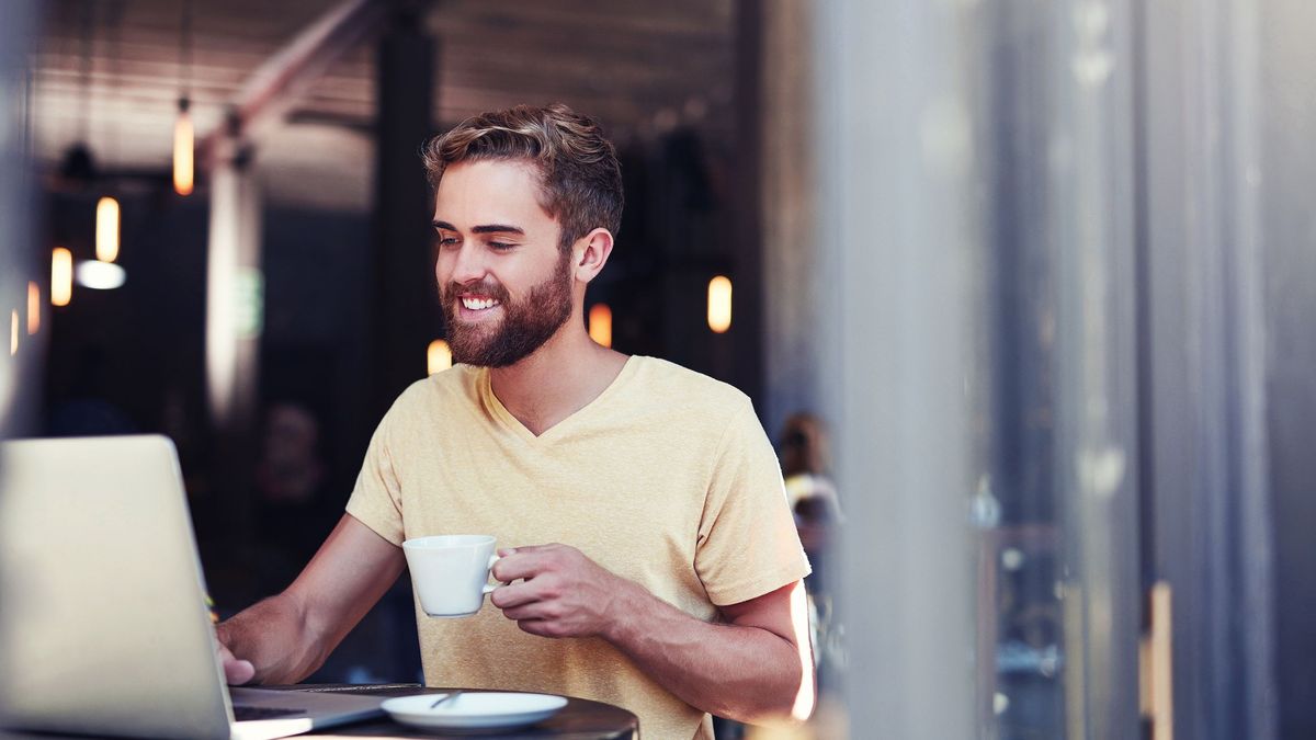 A smiling man sat in a cafe holding a cup of coffee and using a laptop