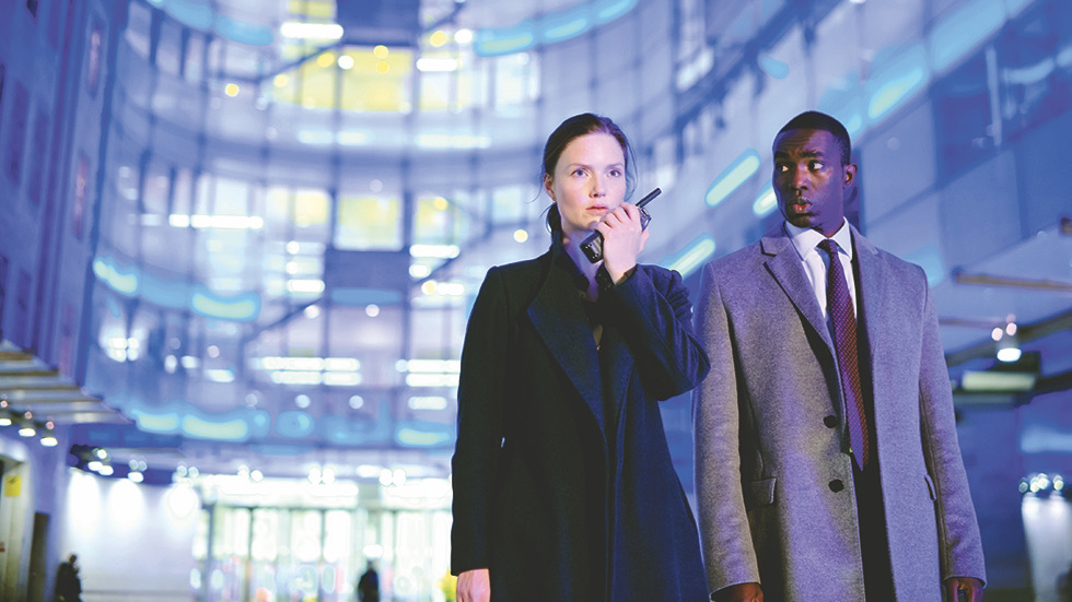 Holliday Grainger dans un pardessus sombre tenant un talkie-walkie comme Rachel Carey et Paapa Essiedu dans un costume et une cravate et un pardessus gris alors qu'Isaac Turner se tient devant la BBC à Londres dans The Capture saison 2.