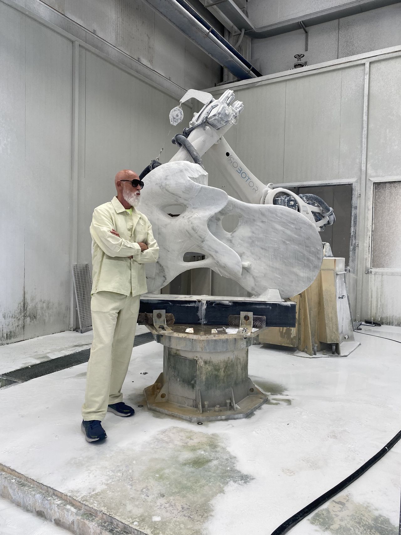 Man dressed in white clothes standing next to marble sculpture in a workshop setting