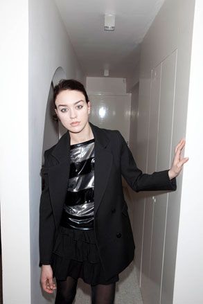 Female model, wearing black blazer and shorts, metal silver and black striped top, white corridor backdrop