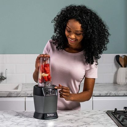 Woman in pink top in the kitchen using the ninja food blender with red fruits and ice in it
