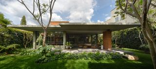 Daytime exterior image of Ventana House in Mexico among greenery, flat white roof, red brick supporting pillar, green lawn, shrubs, plants and trees, windows, stone patio area with irregular shaped stone steps, tables and chairs, blue cloudy sky