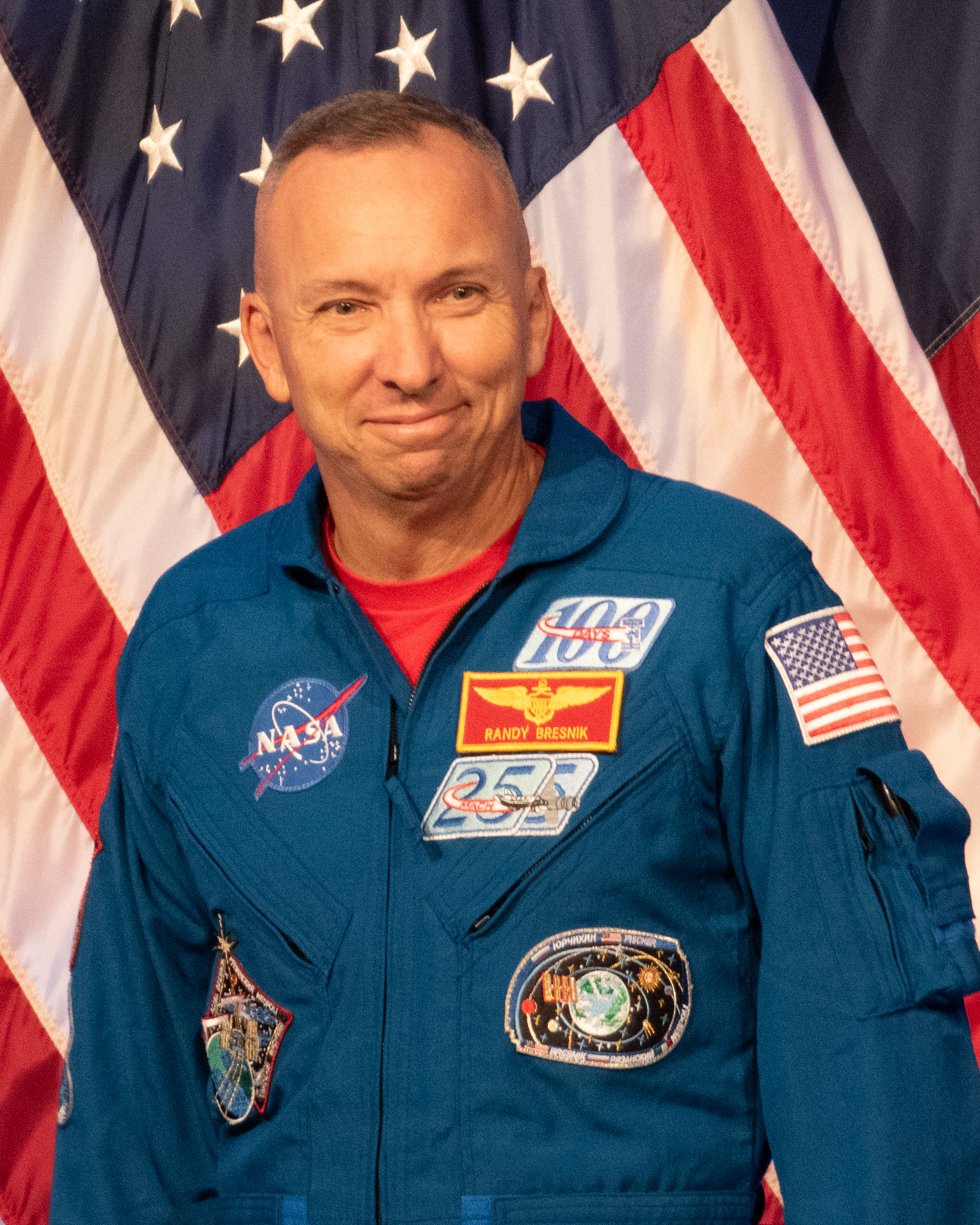 um homem careca de macacão azul sorri com uma bandeira dos EUA pendurada atrás dele.