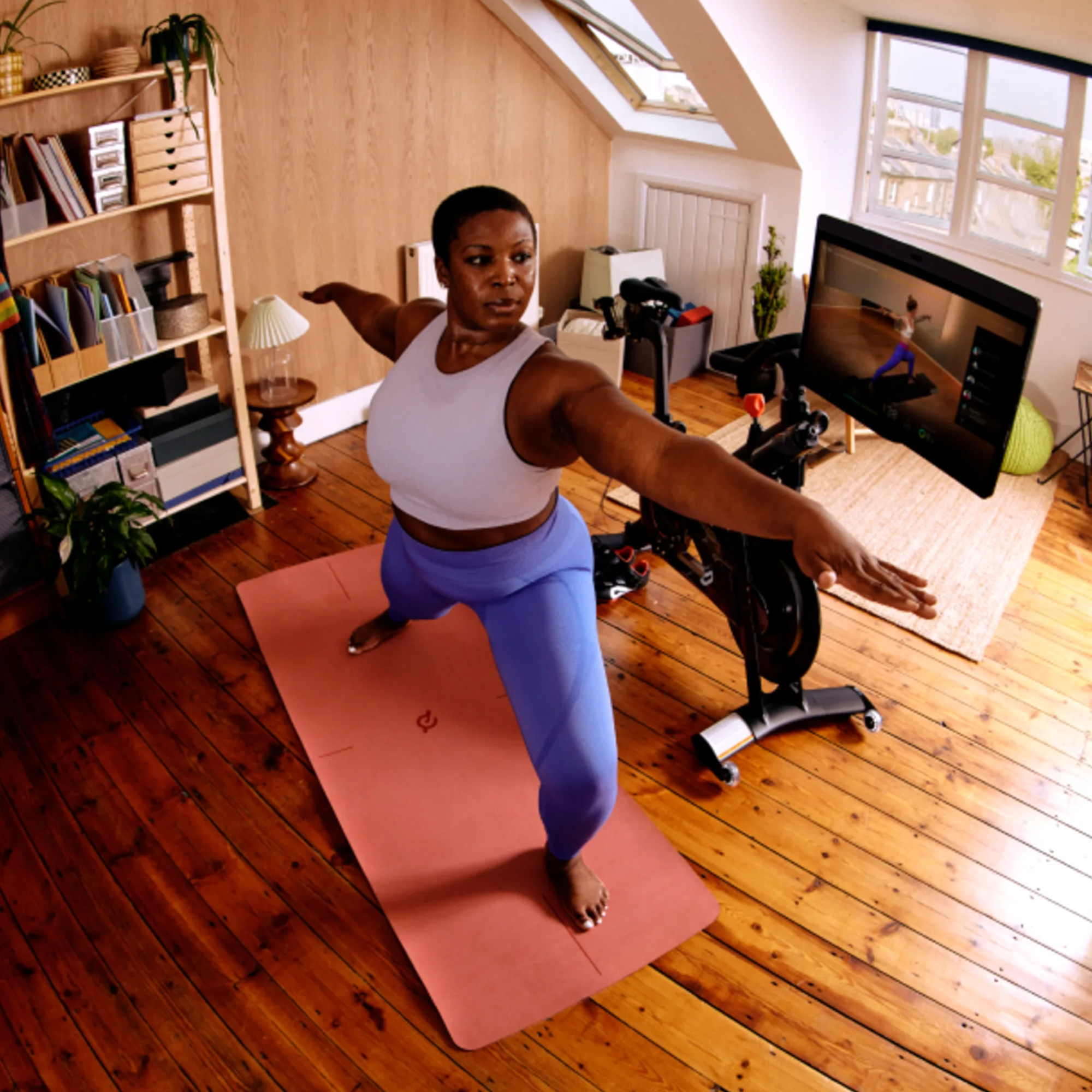 A woman standing on a mat doing exercises in a loft room beside a Peloton exercise bike