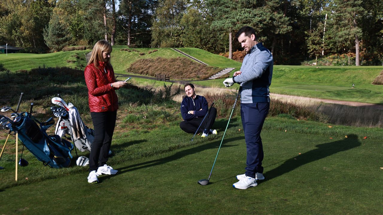 Three golfers waiting on the tee due to slow play, with one on their phone, one sitting on the floor and one checking their watch