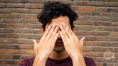 A young man puts his hands over his eyes like he's trying not to look at something.