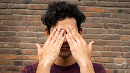 A young man puts his hands over his eyes like he's trying not to look at something.