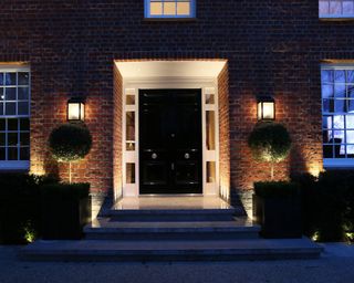 A black front door with spherical shrubbery either side and two wall lamps illuminating the porch