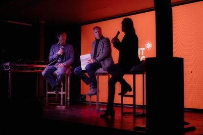 Darkly lit stage, three men sat on stools, orange screen at the back of the stage, far right male and stage column silhouetted