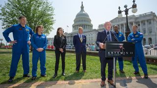 a man in a suit speaks at a lectern outside while four astronauts in blue flight suits and two well-dressed people look on