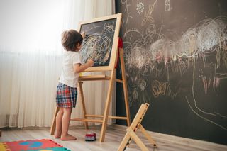 A boy is drawing on a small chalkboard, there is a large chalkboard wall to the right of the picture.