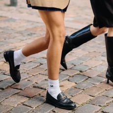 Two women walk along a cobbled street, one wears black loafers with white sports socks and the other black knee-high boots.