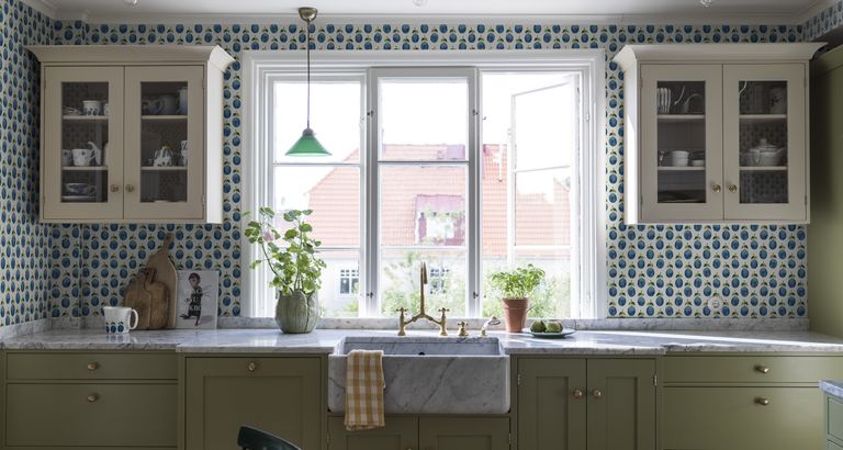 Kitchen wall decor ideas by Borastapeter, with blue and white patterned wallpaper and wall mounted cream cabinets either side of a window.