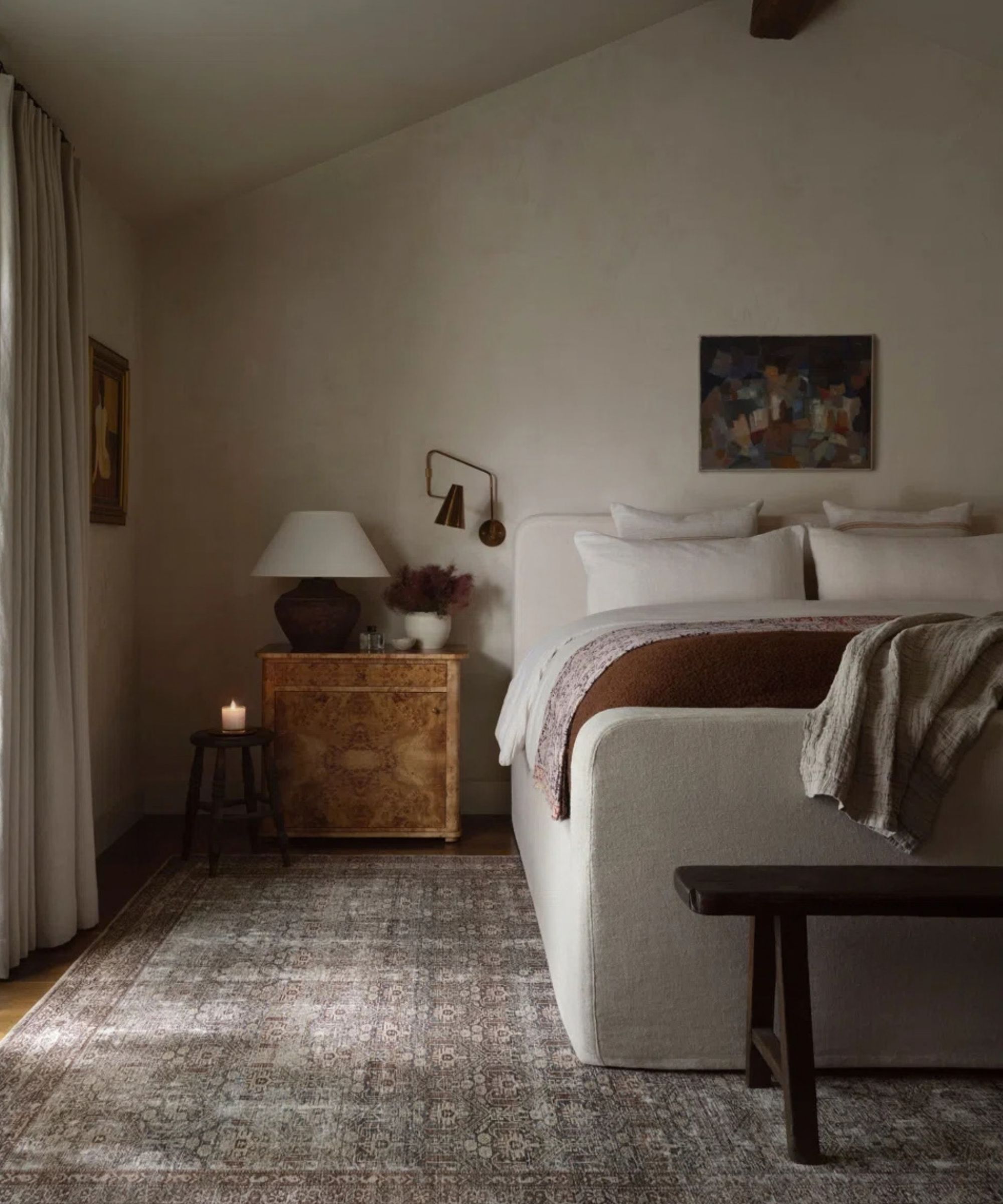 Autumnal bedroom with beige plaster walls, beige bed with a dark orange throw, a burl wood nightstand, a large brown Persian-style rug, and a side table with a candle on top.