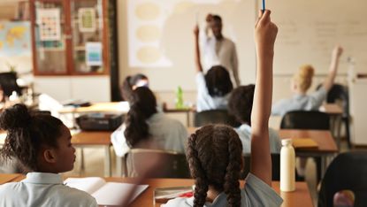 Kids in a classroom, some with their hands raised to answer a question.