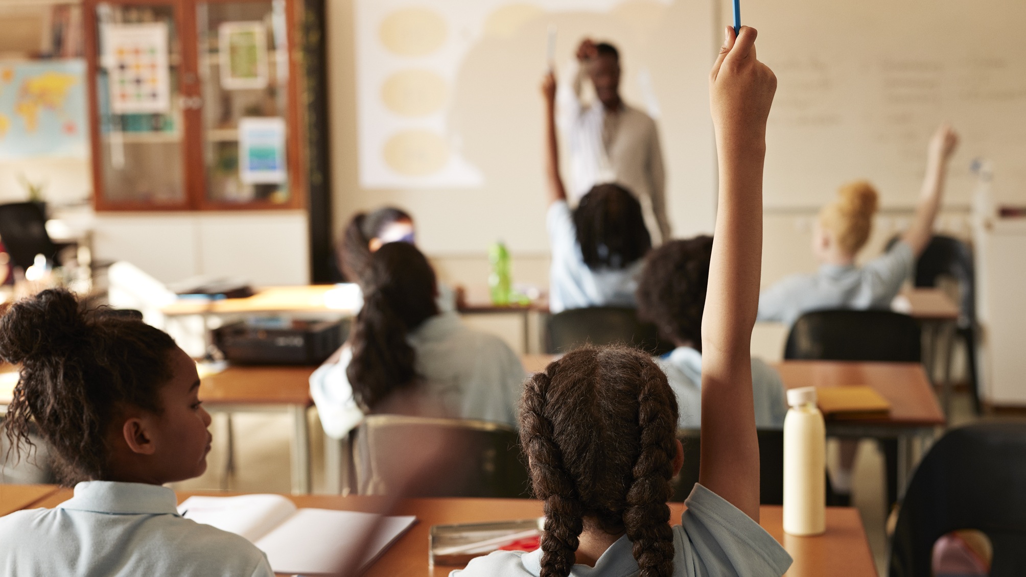 Kids in a classroom, some with their hands raised to answer a question.