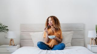 A woman sat crossed legged on a bed eating a bowl of food and smiling