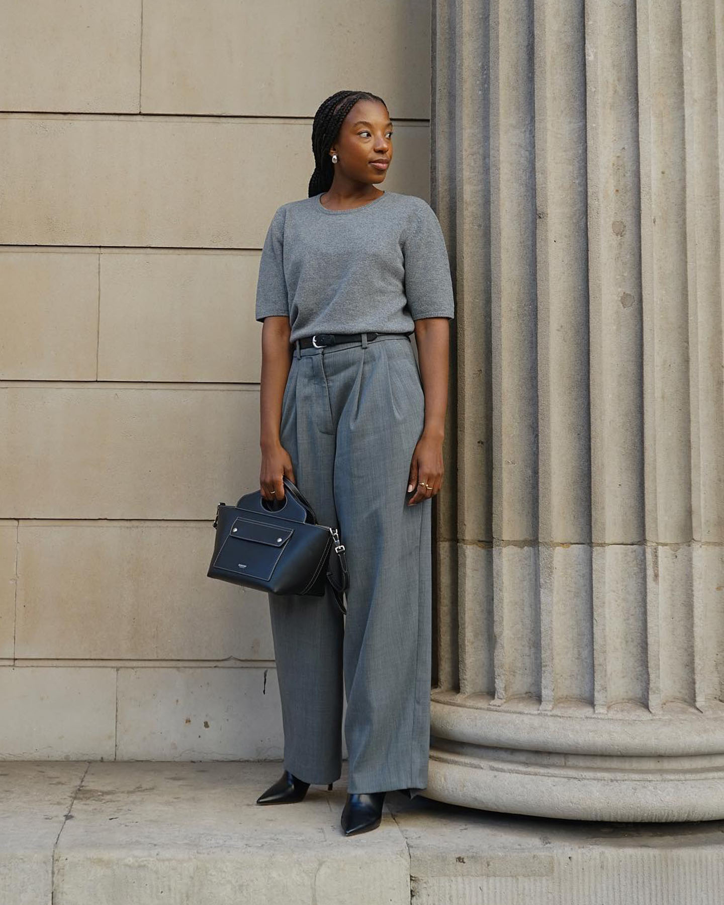 British influencer wears an all-gray outfit witj a short-sleeved sweater, belted trousers, a black bag, and black pointed-toe boots.