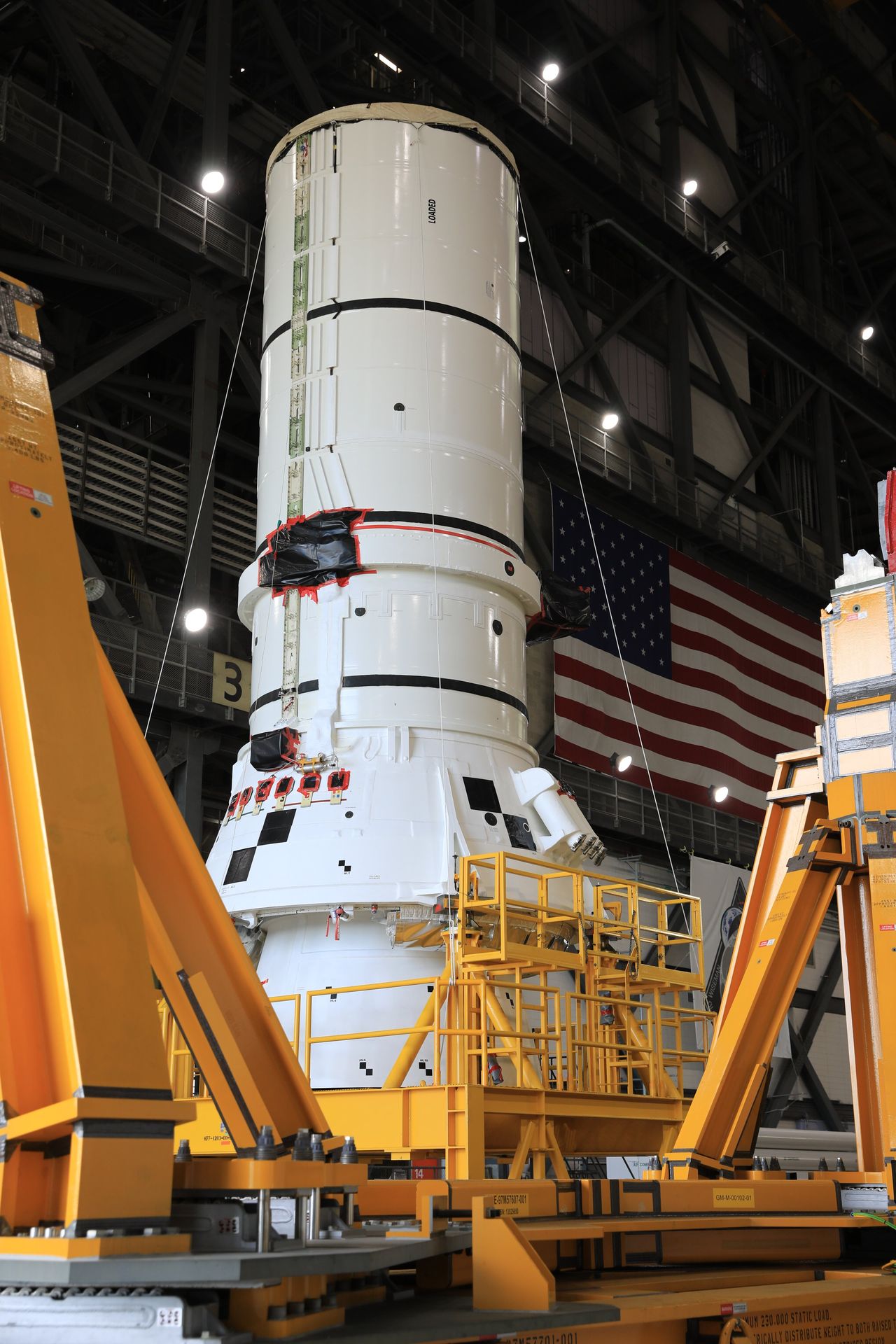 The bottom segment of a white solid rocket booster stands on a yellow support inside a giant rocket hangar.