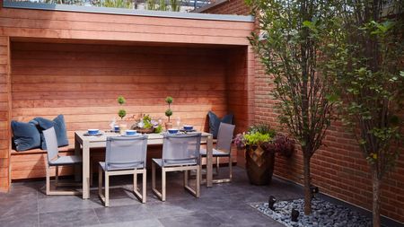 Backyard with grey floor tiles, wood panelling, banquetting area, grey chairs, wooden rectangular table, grey pillows, table set, planter, 