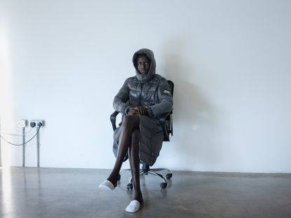 Man sits in chair in Moncler grey puffer jacket in concrete room with white wall
