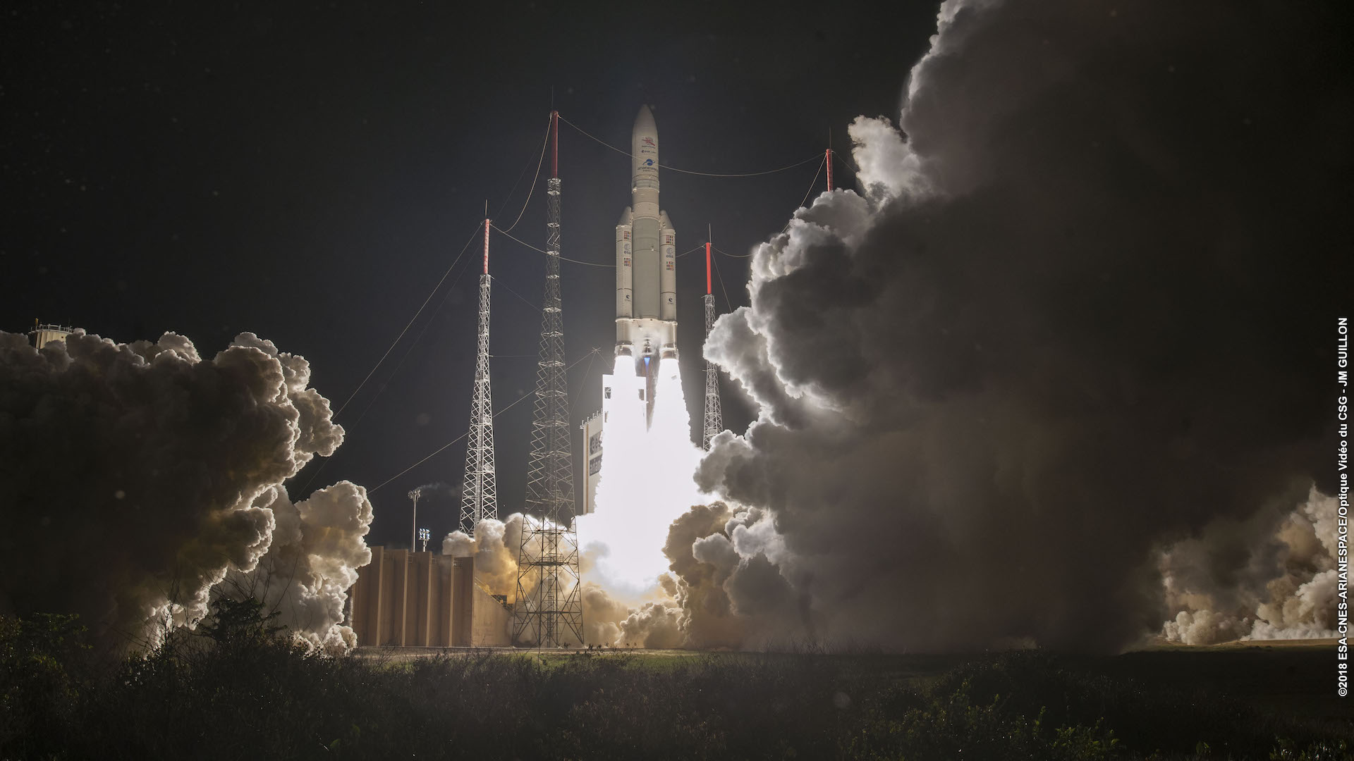 An Ariane 5 rocket lifts off in the dark of night, as its two side booster blast a blinding white fire, a cloud of smoke rises from the launchpad