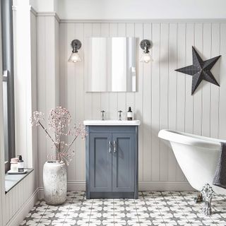 bathroom with white panelled wall, grey and white star pattern floooring, a freestanding white rolltop clawfoot bathtub, with a blue vanity and two wall lights