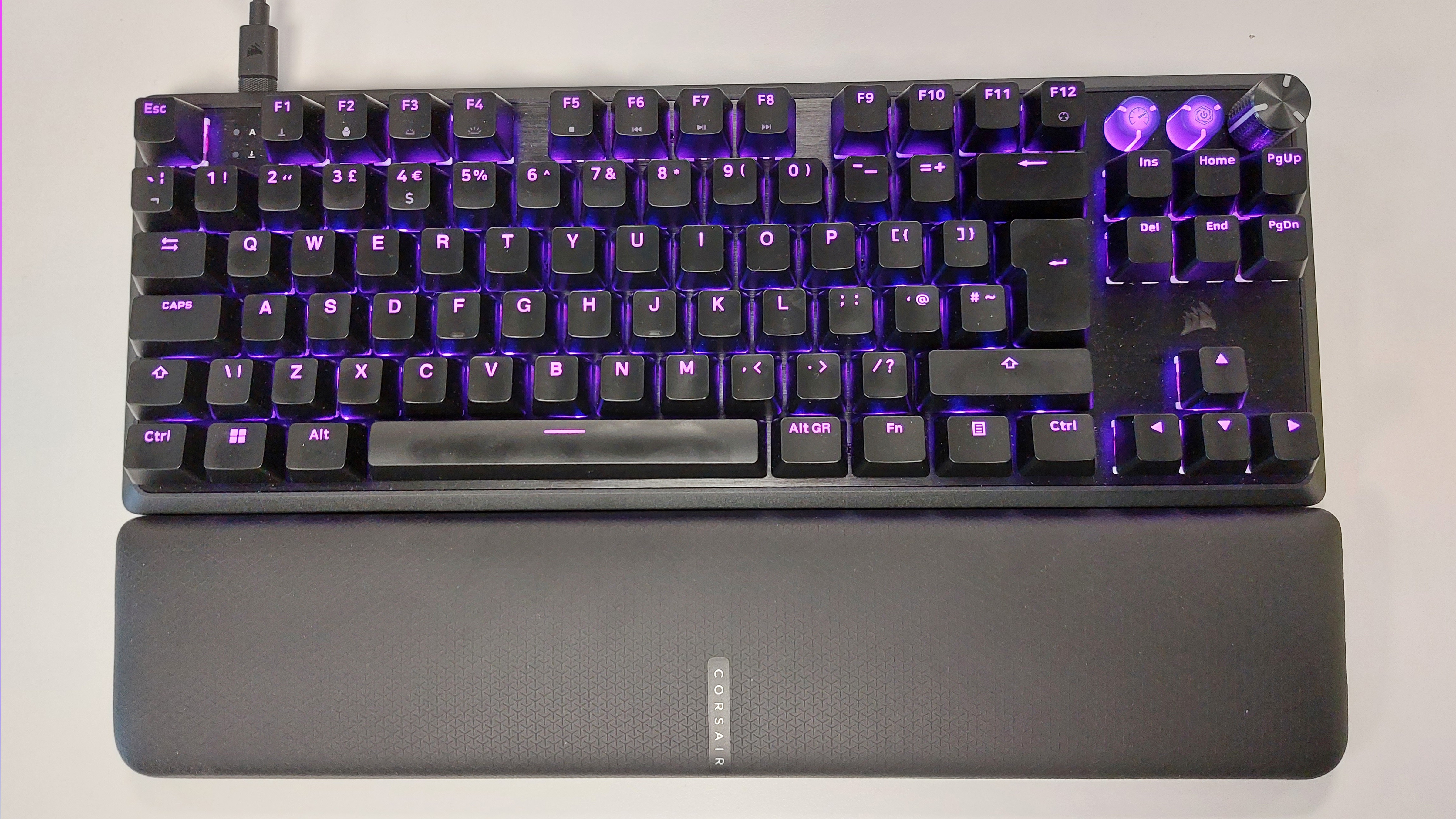 The Corsair K70 Pro TKL gaming keyboard and magnetically attached memory foam leatherette wrist rest seen from above on a well lit white desk. The keyboard is plugged in, with the RGB lights powered on. All of the keys are bathed in a purple light.