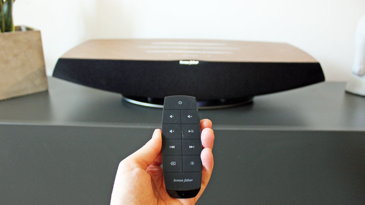The remote control with the sonus faber omnia in a man's hand, being pointed at the speaker. The remote has playback and input controls in large buttons that cover its whole area.