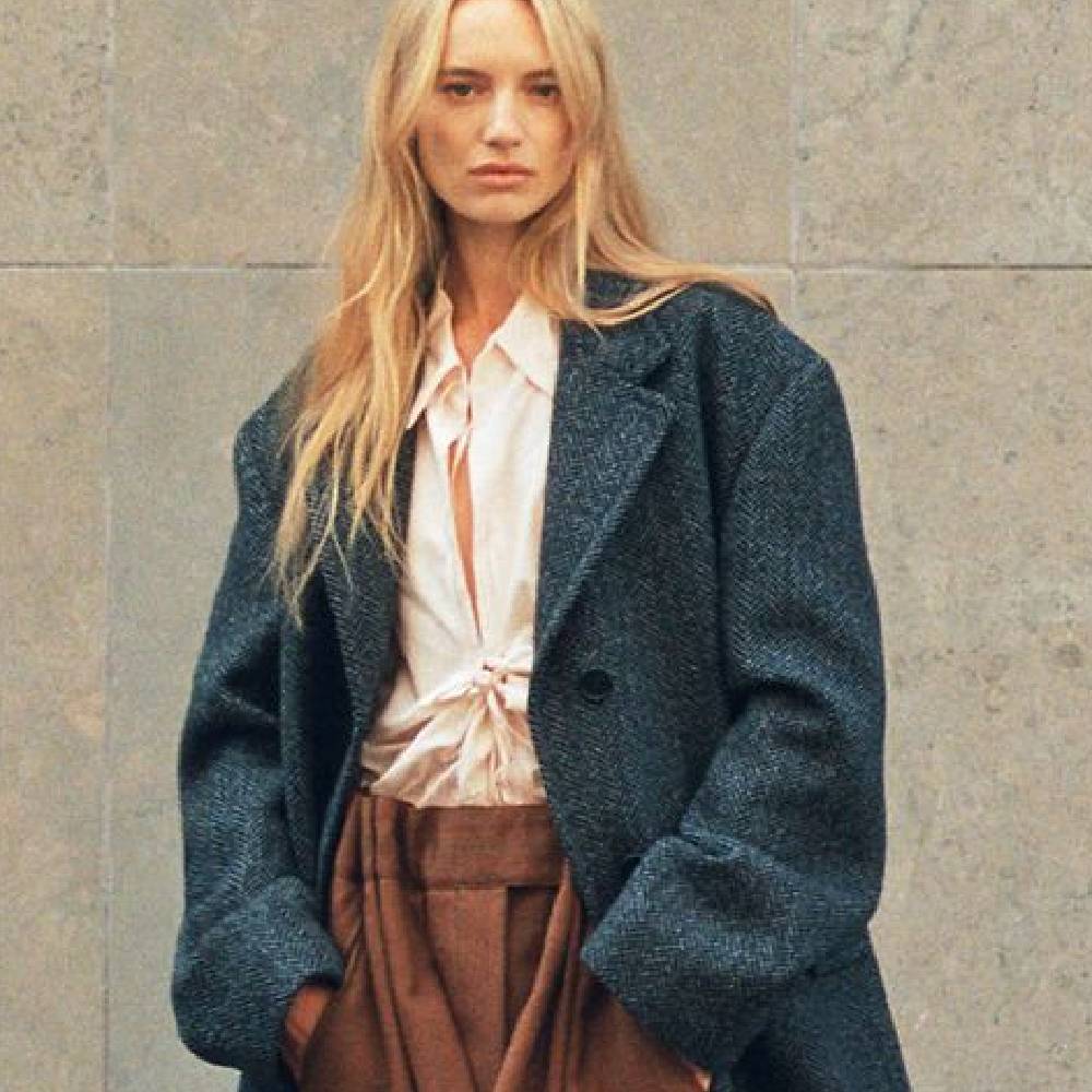 A female model wearing a cream blouse, brown suit trousers and a dark blue/grey coat from Farfetch.
