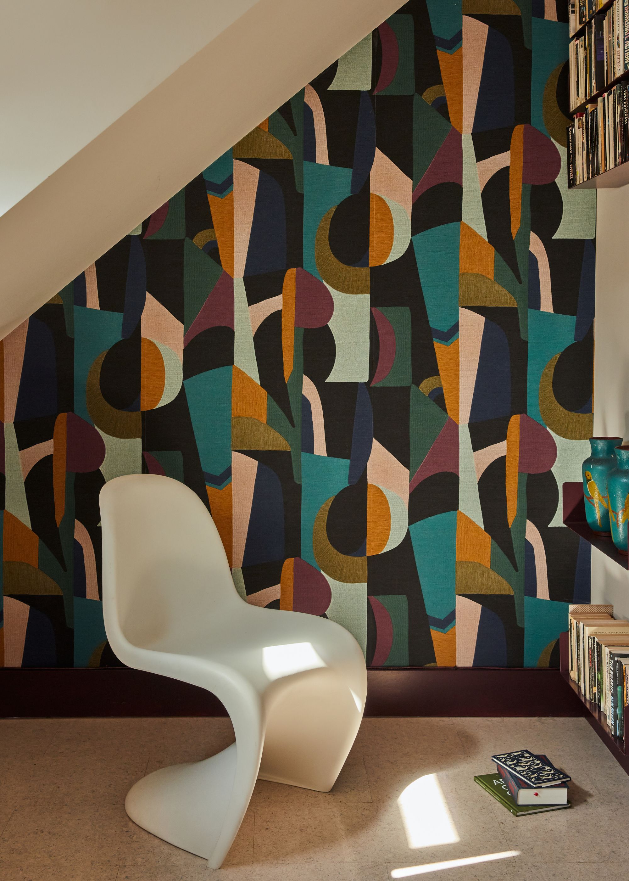A corner of a room with a sloped ceiling, bookshelves, and a white modern chair. The wall is lined with a colorful, abstract print wallpaper.