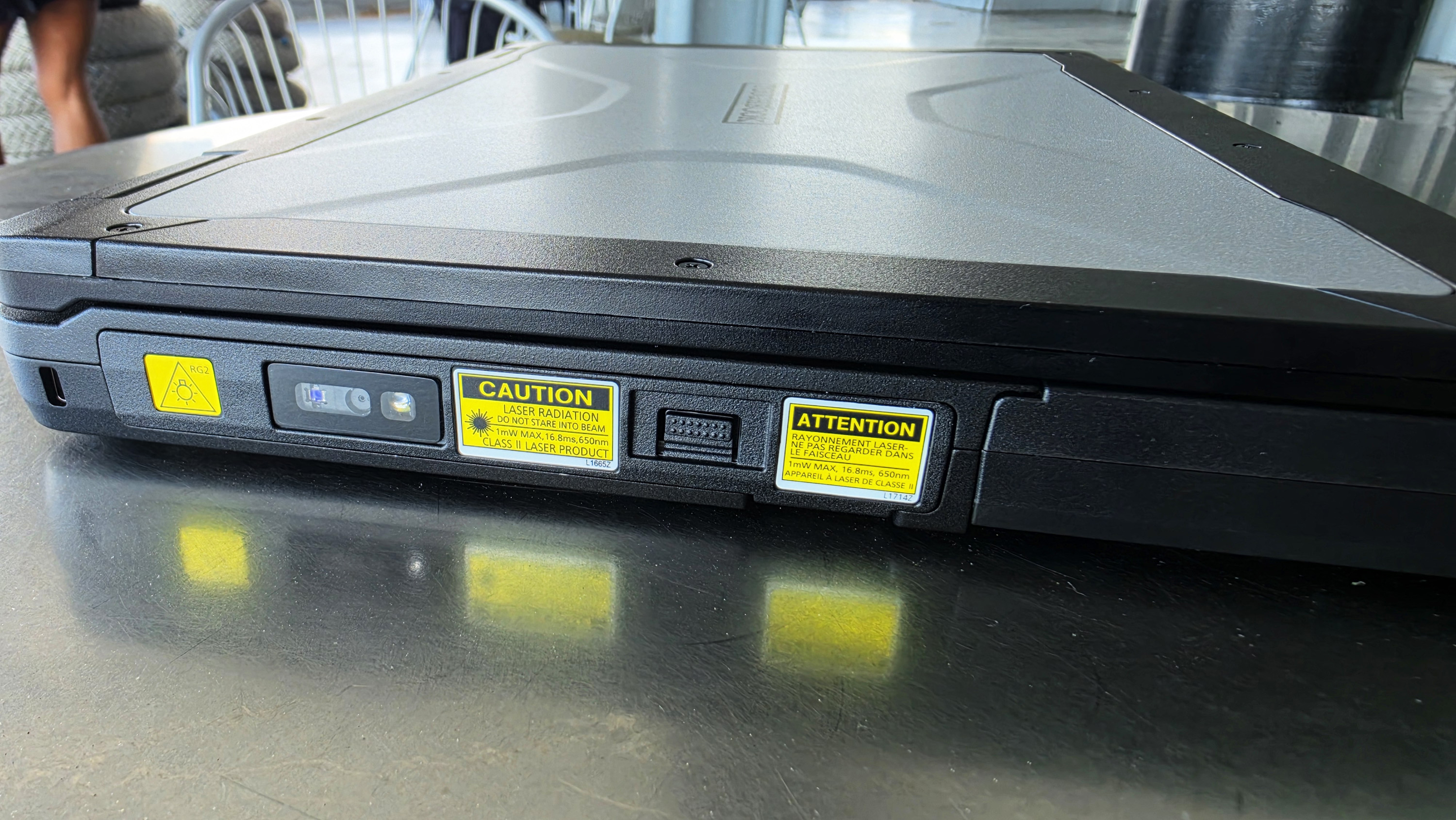 A rugged laptop on a reflective metal table, showing its closed lid. Yellow caution labels warn of laser and electric hazards, emphasizing durability and safety.