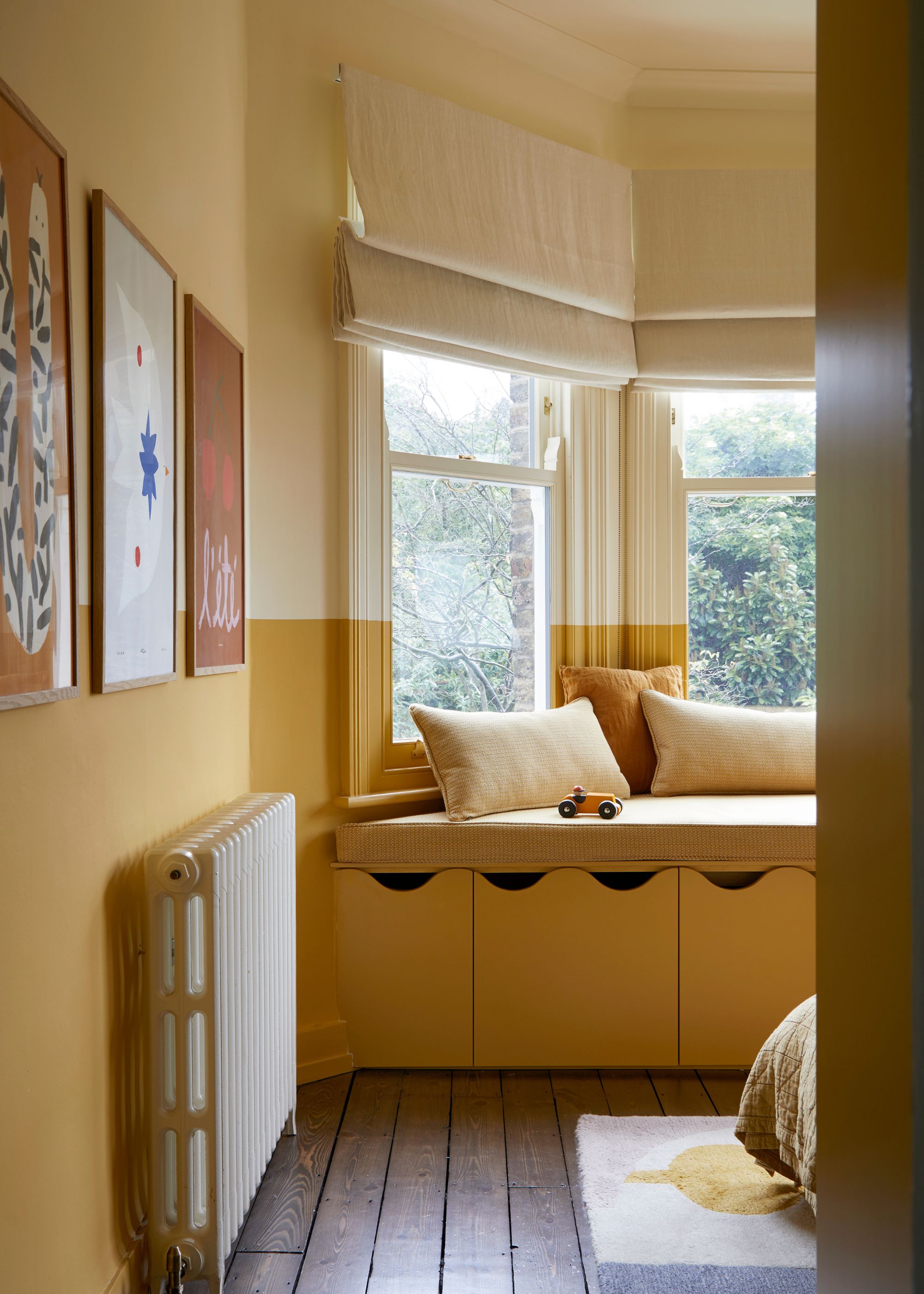 A bedroom with dark yellow walls on the lower half, cream walls on the upper half. A bay window with a window seat, cream Roman blinds, and neutral cushions.