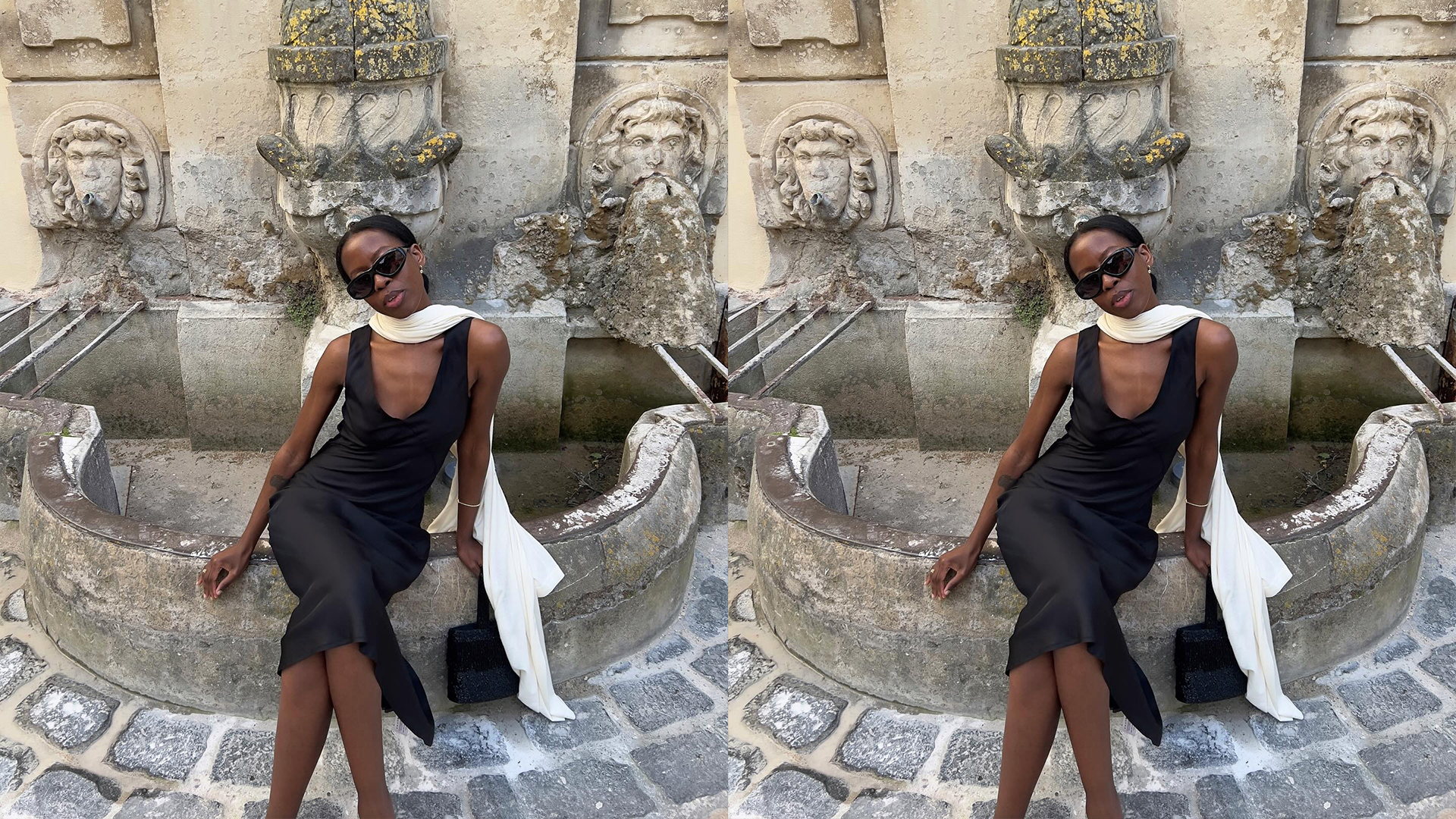 Sylvie wears black dress and white scarf while sitting on a fountain and posing for the camera. 