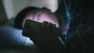 A man lies in bed looking at his phone, which shines a bright light into his face