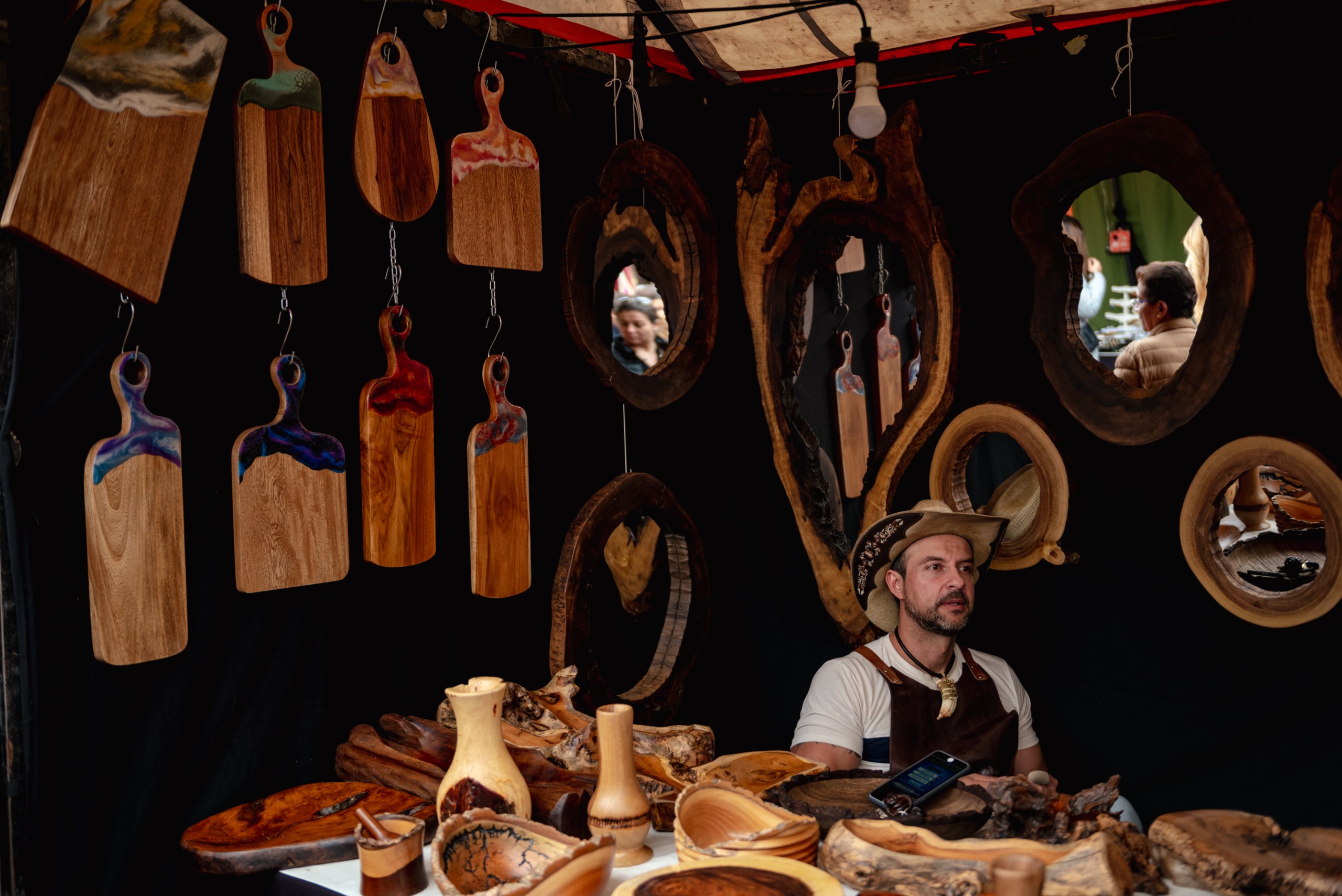A street market vendor sat in his booth wears a hay cowboy hat surrounded by wooden creations such as chopping boards, mirrors, and vases.