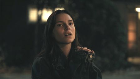 Nikki in a jacket with her hands together in Obsession. 