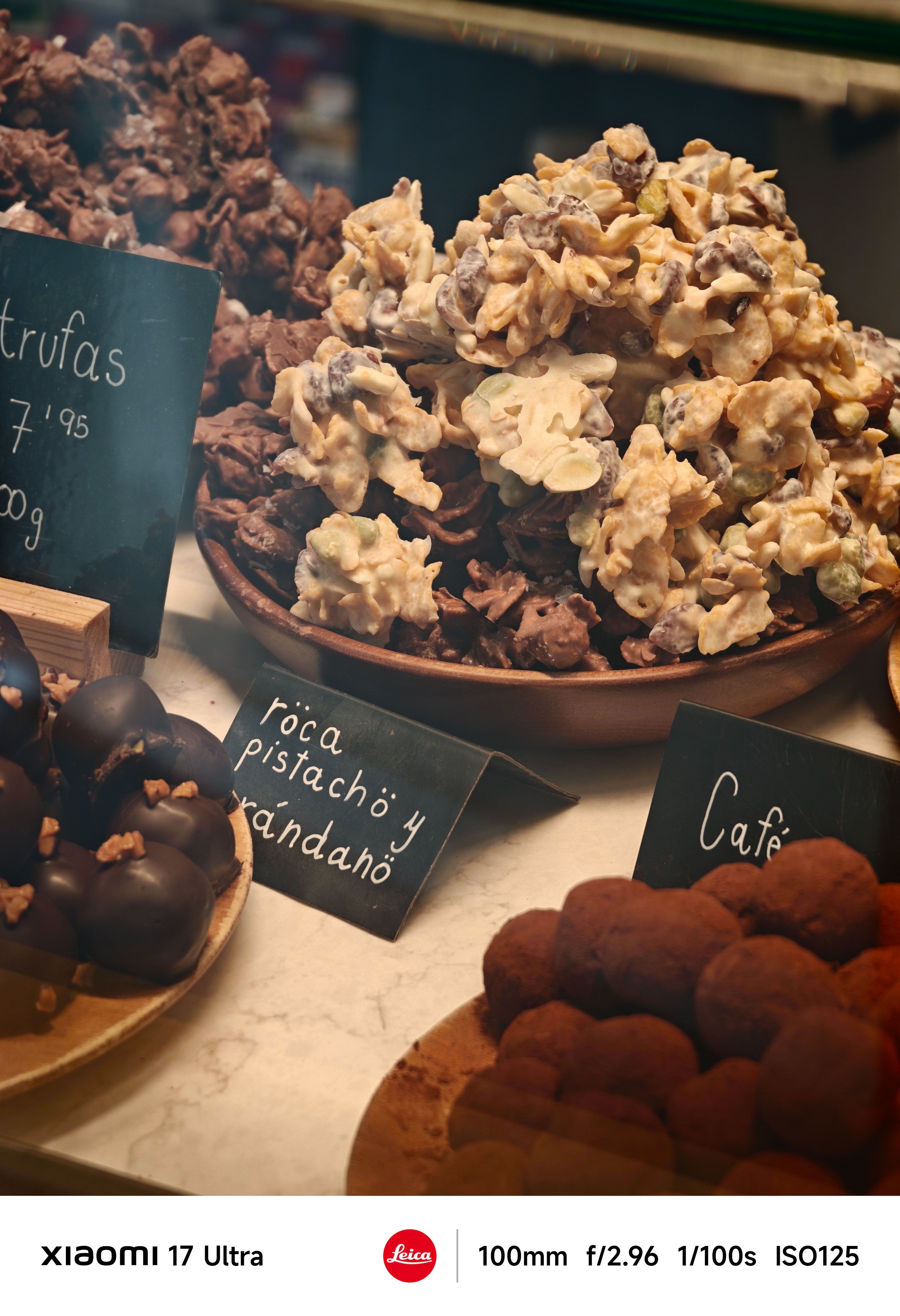 Tight telephoto shot of chocolate truffles and caramelized nut clusters in a bowl, shallow depth of field with shop labels partially visible.