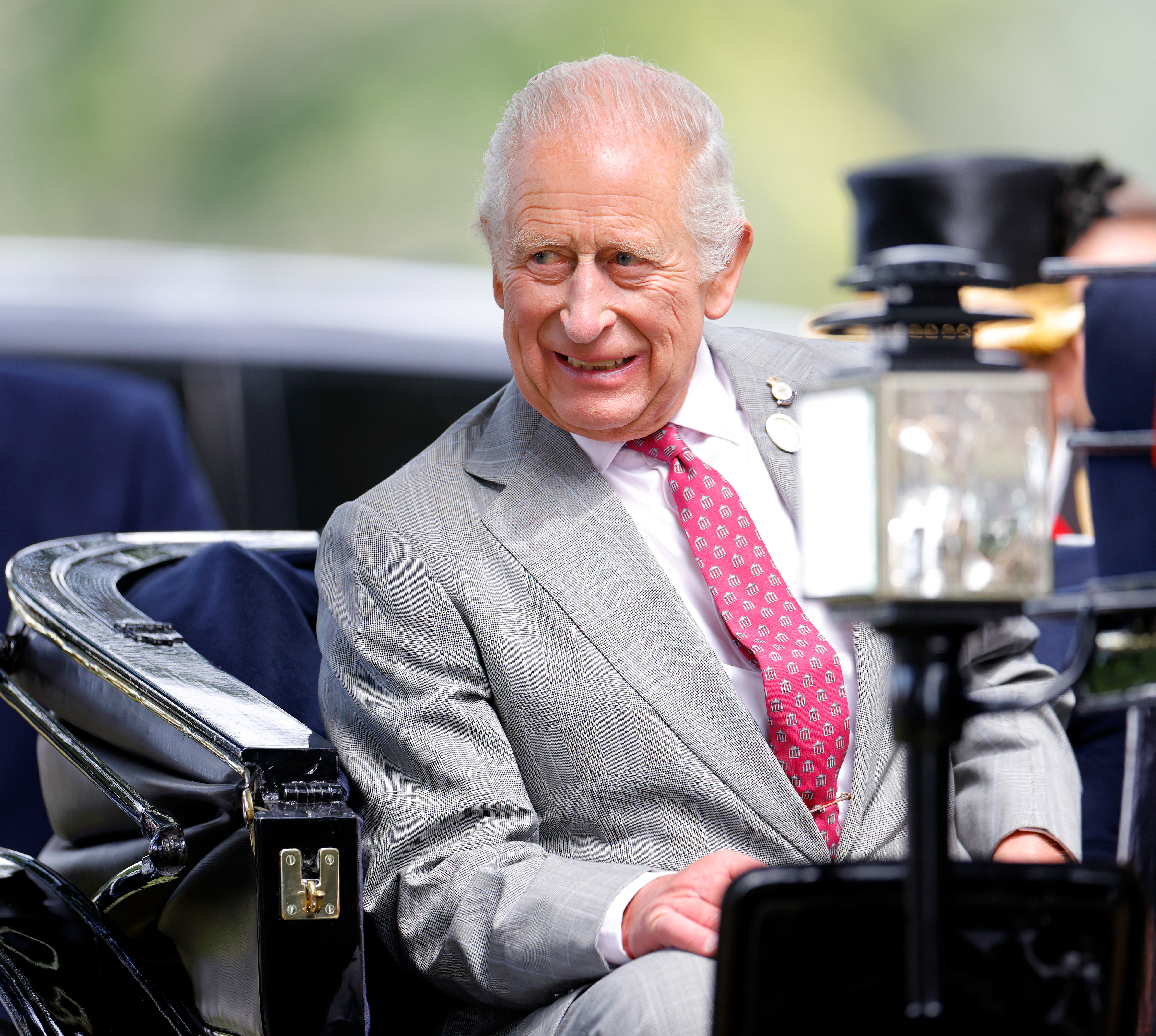 King charles wearing a gray suit and red tie sitting in a carriage and smiling