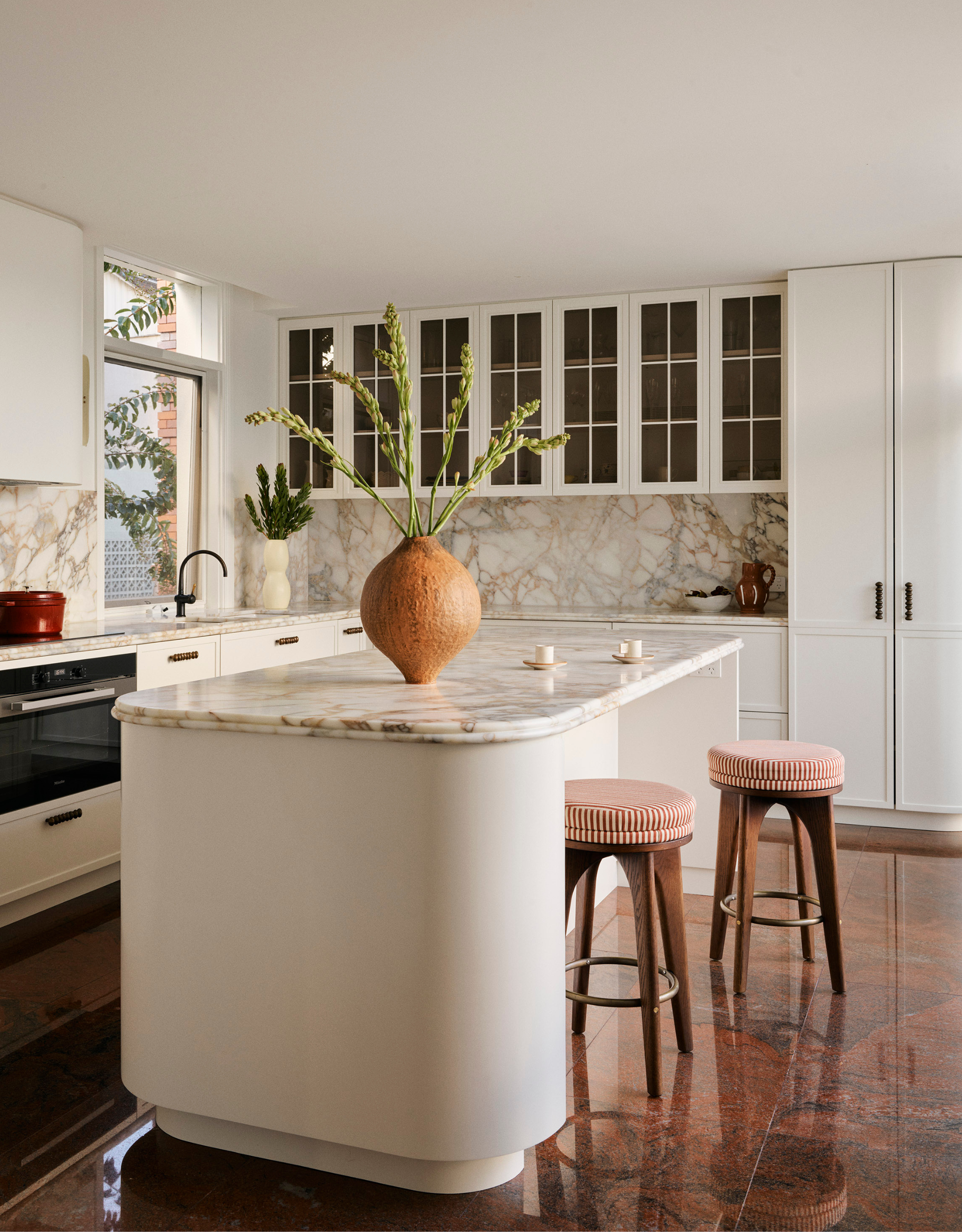 A bright white kitchen with a stone backsplash, ribbed glass cabinets, a curved island, a pair of striped stools, and a vase with flowers