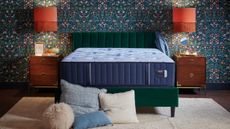 Stearns & Foster Estate Mattress on a dark velvet bedframe in a bedroom with blue floral wallpaper, orange lamps, and matching dark wood bedside tables