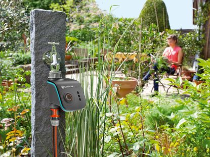 best self watering gadgets and accessories, garden and greenery with a smart irrigation system connected to an outdoor water tap and a woman sitting in the background 