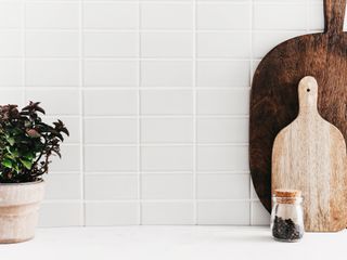 White kitchen tiles background with two wooden decorative chopping boards, a plant in a pot, and a jar with peppercorn inside.