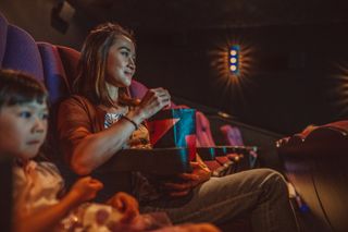 Young Asian mom & her lovely daughter eating popcorn while watching movie in movie theatre.
