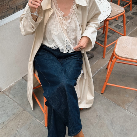 Fashion person Chloe Kath Butler wears a transitional winter to spring outfits 2026. She wears a embroidered blouse, blue jeans and brown boots. She has a cream trench over her shoulders. She is sat down, looking to the side drinking prosecco in a recent image from her Instagram. 