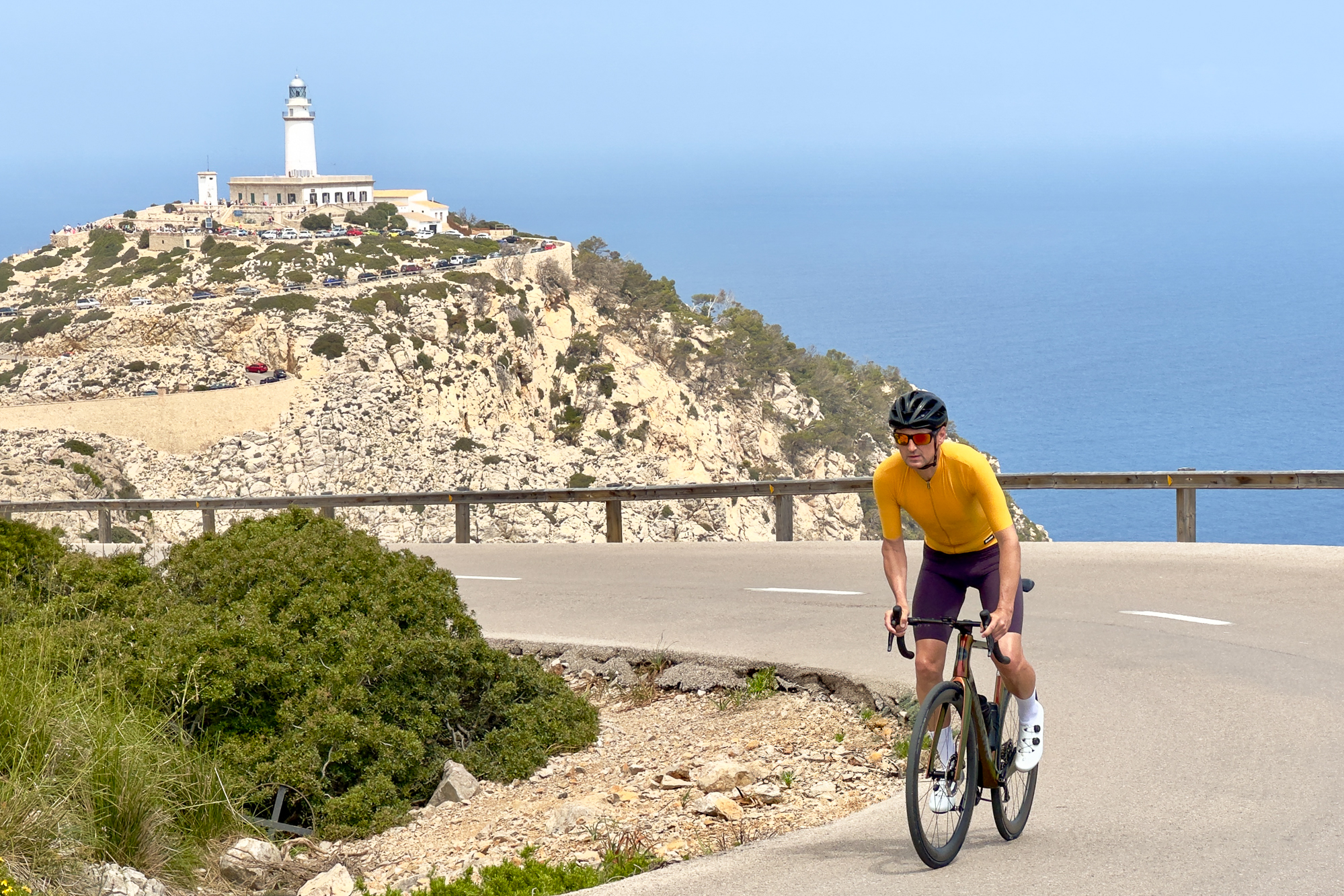 Man wearing a yellow jersey riding out of the saddle on a Handsling bike at Formentor, Mallorca, with a lighthouse in the background