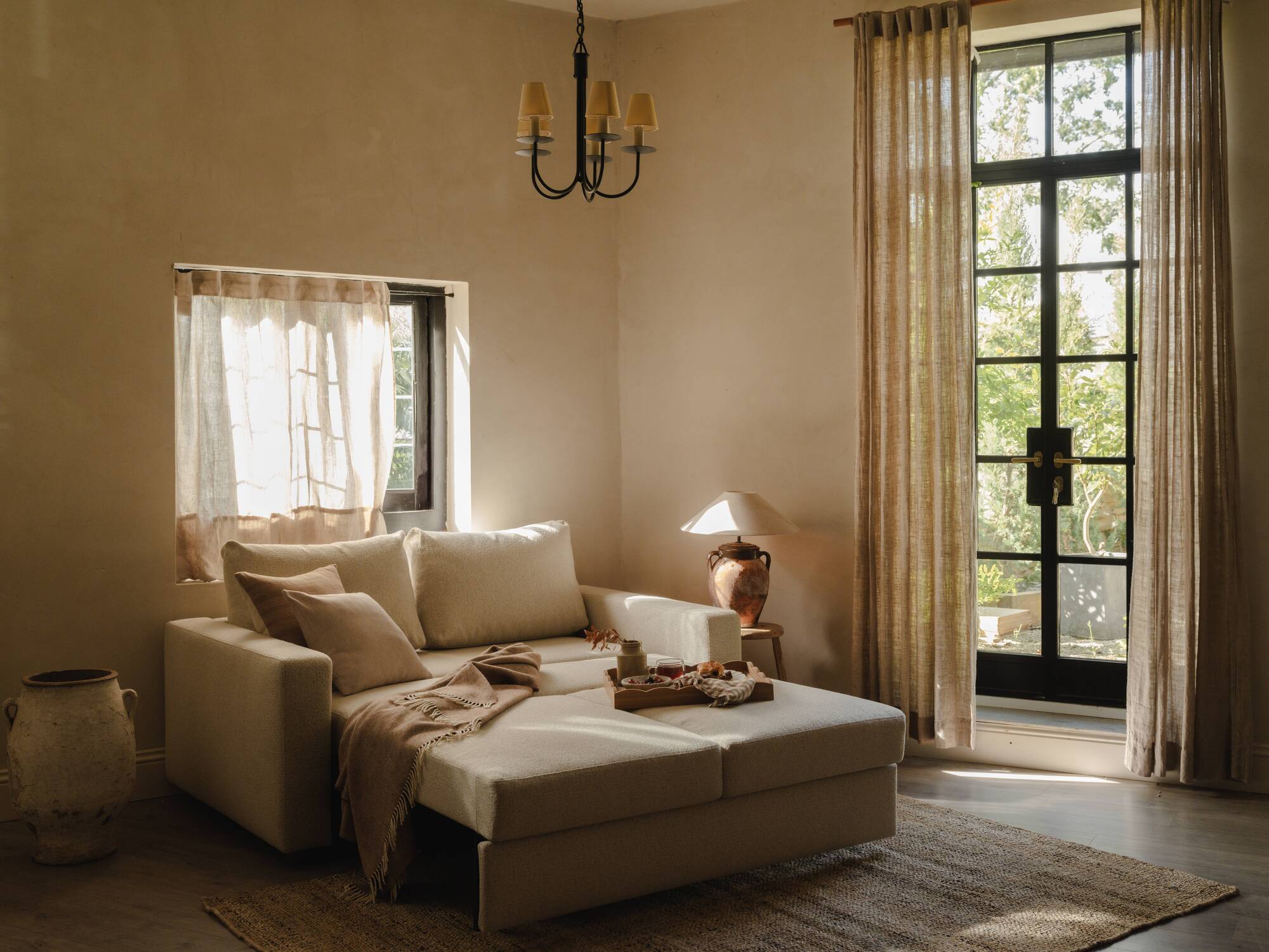 modern room with beige walls with a folded out sofa bed styled with throws and cushions, a window above with sheer curtains, a steel-framed door with sheer curtains, a chandelier, and side table with a lamp