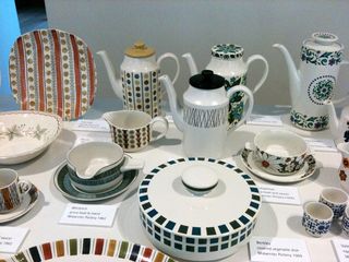 Decorative tableware - plates, jugs, mugs and bowls - on display. photographed on a white surface, with descriptive tags placed next to them.